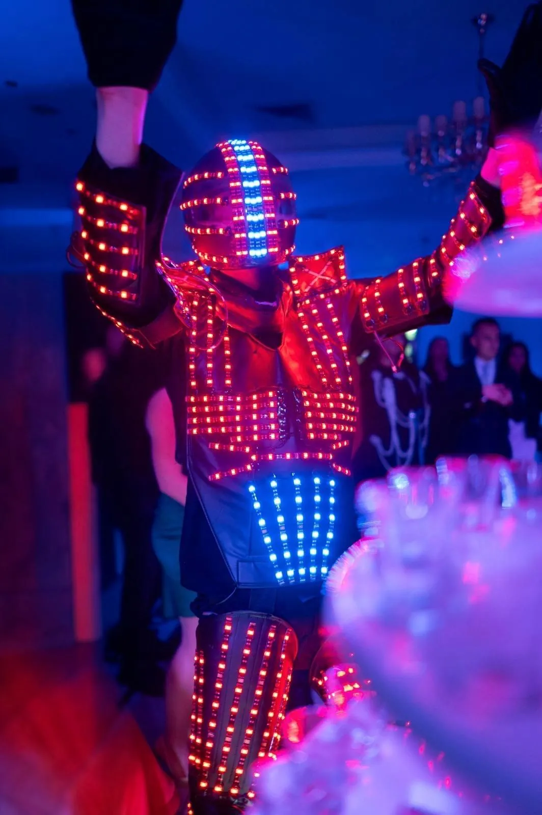 Robotul LED cu brațele ridicate, în lumini albastre și roșii, lângă turnul de shoturi